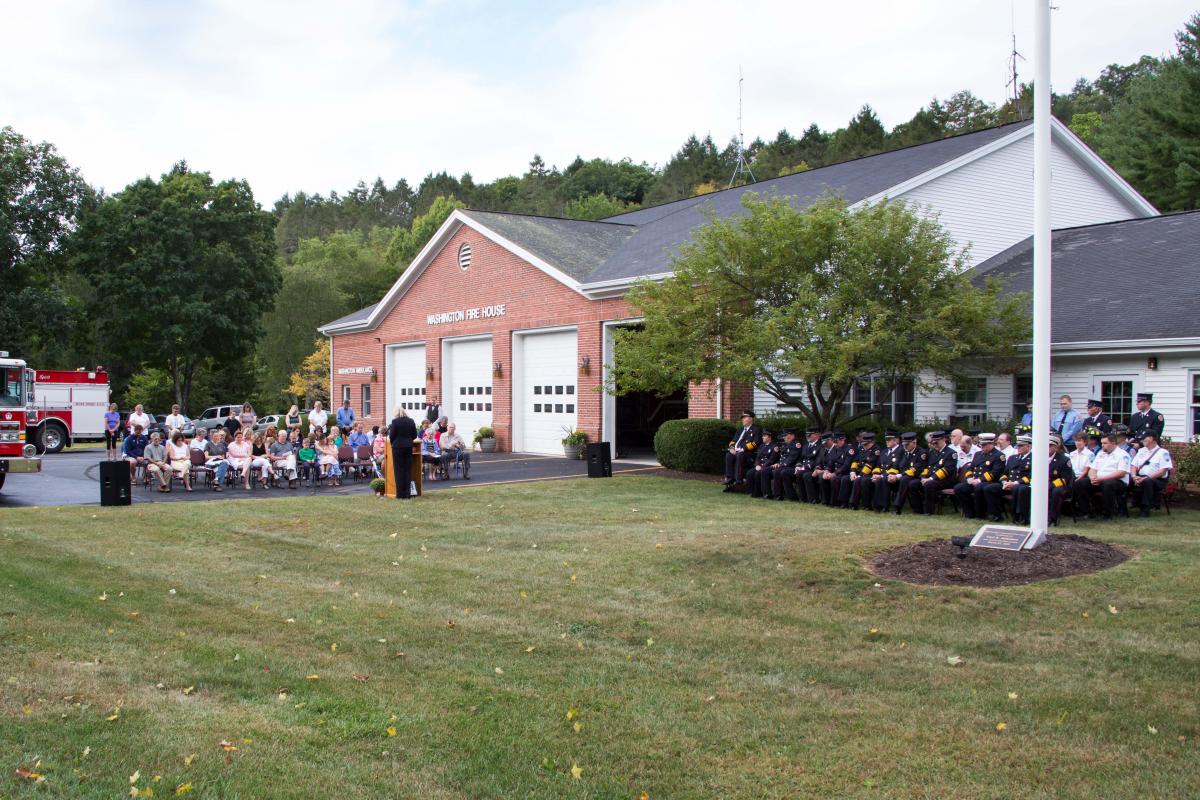 9/11/2016 Ceremony - fire house in center, people sitting in Firehouse parking lot on left, firefighters seated on right on gras