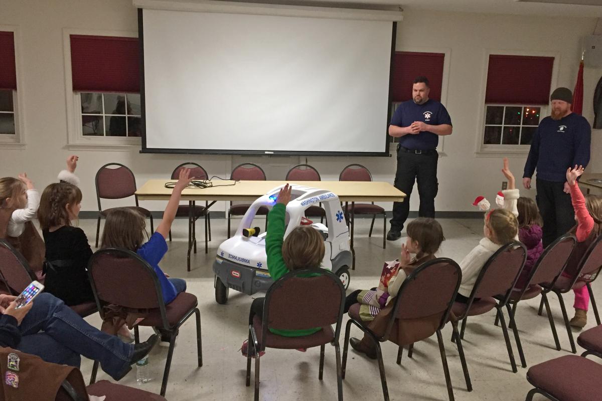 Brownie Troop #40226 visit 2016 -young girls sitting in chairs facing mini-ambulance and firefighter standing in front of screen