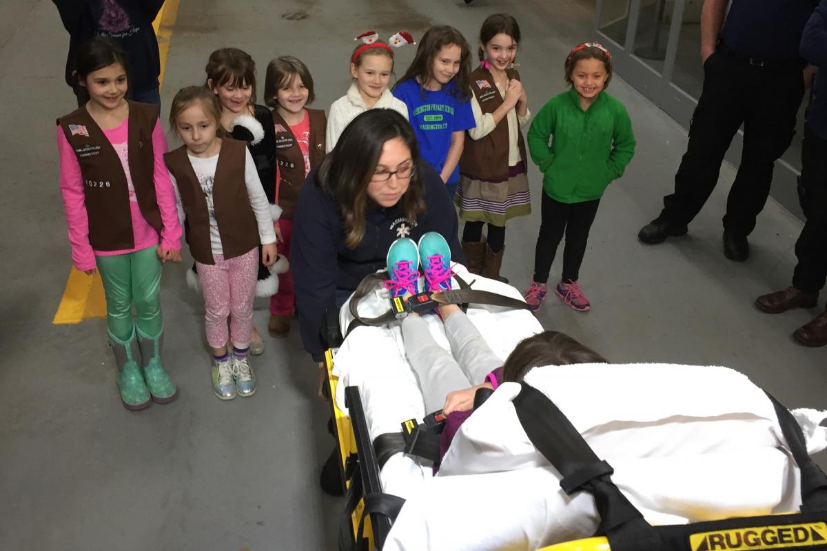 Brownie Troop #40226 visit 2016 - young girl pretending to be patient on gurney, group watching, leader attending to her