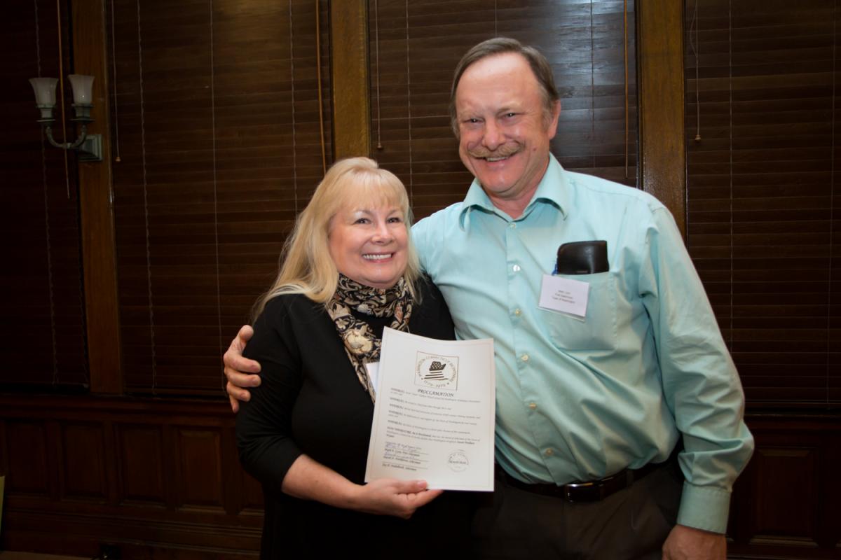 Reception honoring Susie 1/29/2016 - Smiling Man standing with arm around smiling woman who's holding a paper facing camera