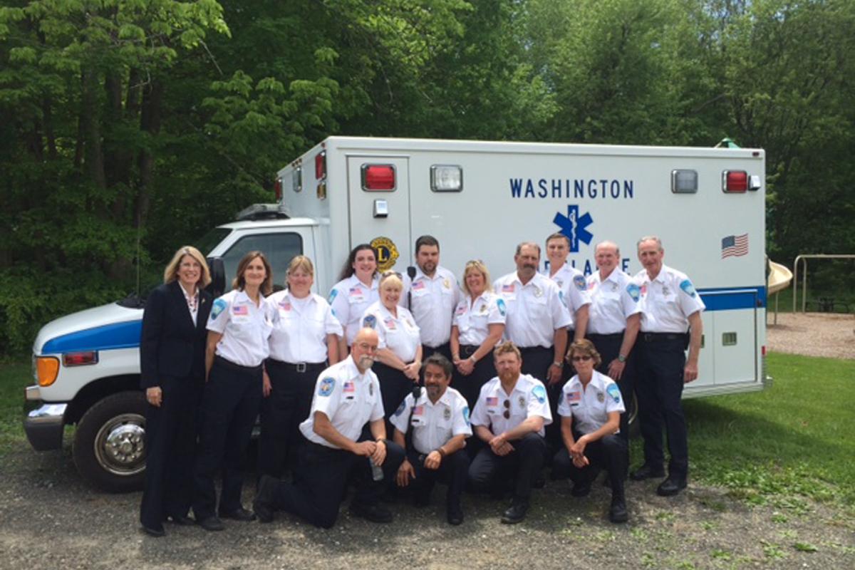 Group at Memorial Day Parade 2014 - 2 rows of people, 1 kneeling, 1 standing along side of modern ambulance, trees in background